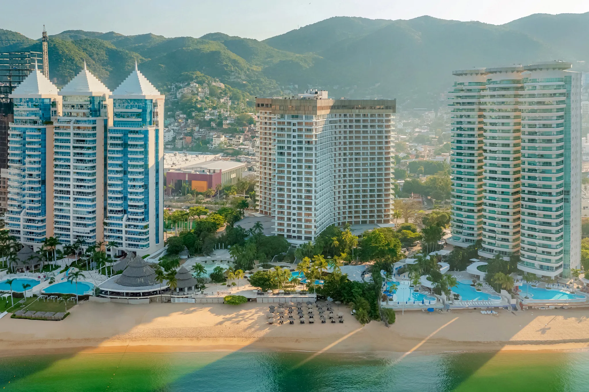 Aerial view of Dreams Acapulco Resort & Spa set along the golden sands of Acapulco Bay with Sierra Madre mountains in the background.